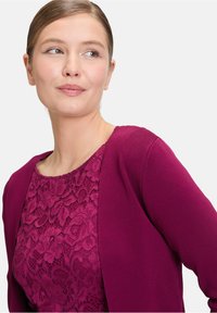 Cardigan fuchsia sur un haut en dentelle de teinte assortie. Tissu doux et texturé avec un col rond et une fermeture ouverte, mettant en valeur le design superposé.