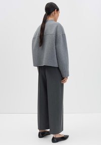 Femme avec une longue queue de cheval noire portant une veste grise ample, un pantalon large gris foncé et des ballerines noires, se tenant devant un fond uni.