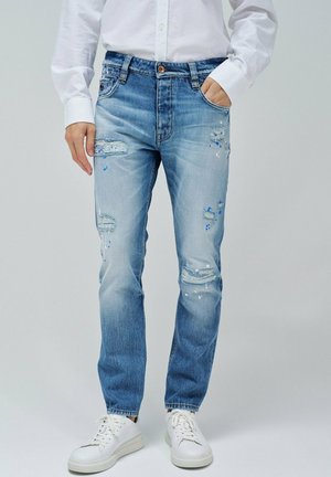 Jeans de mezclilla azul claro con parches desgastados y manchas de pintura azul. Diseñados con un corte recto y el estilo estándar de cinco bolsillos.