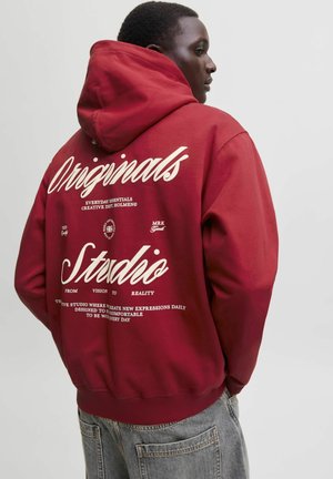 Ung mand iført en rød hættetrøje med hvid tekst ""Originals Studio"" og yderligere små bogstaver på ryggen, kombineret med grå jeans.