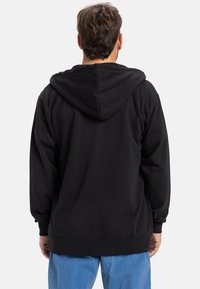 Sweatshirt preto com capuz, de mangas longas e corte solto. Apresenta uma textura suave e punhos canelados, combinado com calças azuis.
