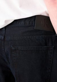 Tamsios džinsinės kelnės su glotnia siluetu, su odiniu Calvin Klein etiketė ant nugarinės juostos ir standartiniu užpakaliniu kišenės dizainu.