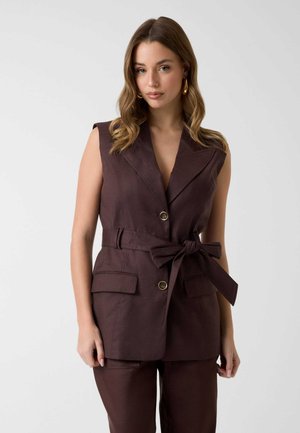 Mujer con chaleco sin mangas marrón oscuro con cinturón y botones y pantalones a juego, de pie frente a un fondo claro y liso.