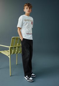 Tricou alb cu grafică, având un model în carouri și o inimă roșie, asortat cu blugi negri și adidași alb-negru. Scaun verde în fundal.