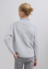 Suéter gris claro de punto con un patrón texturizado de diamantes, cuello con volantes y puños acanalados. Se lleva con jeans grises.