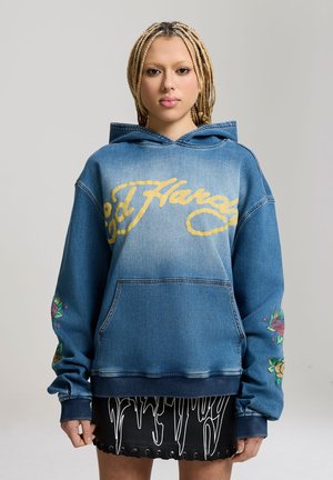 Joven con el pelo trenzado que lleva una sudadera con capucha azul de Ed Hardy con diseños florales en las mangas y una falda negra con patrones gráficos blancos.