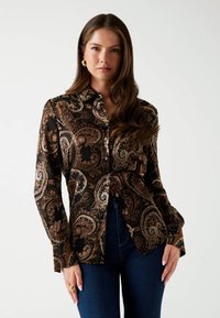 Donna con lunghi capelli castani che indossa orecchini a cerchio d'oro, una camicia a bottoni nera con motivo paisley e jeans blu scuro su uno sfondo bianco.