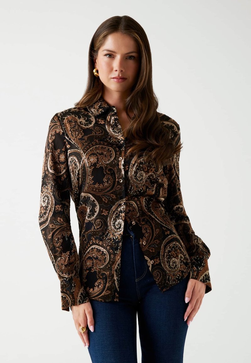 Donna con lunghi capelli castani che indossa orecchini a cerchio d'oro, una camicia a bottoni nera con motivo paisley e jeans blu scuro su uno sfondo bianco.