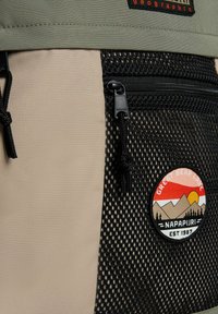 Zaino realizzato in materiali a rete beige e neri, con una tasca con zip e una toppa circolare con un design di montagna e tramonto.
