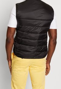 Una persona con un gilet imbottito nero e pantaloni gialli è vista da dietro, mostrando il design trapuntato del gilet e l'outfit casual.