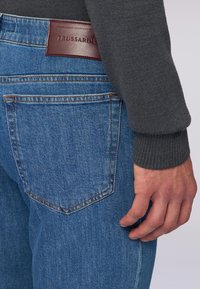 Niebieskie dżinsy z brązową skórzaną metką; mają klasyczne kieszenie i lekko wyblakłą teksturę. Widać mankiet szarego swetra.