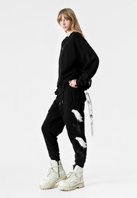 Sudadera negra de gran tamaño con un logo en el frente. Pantalones jogger negros con bordado de plumas blancas y una correa lateral. Un par de botas blancas gruesas.