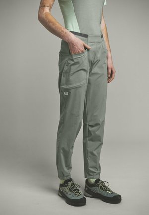 Person trägt hellgrüne Hose mit großen Seitentaschen, hellgrüne Socken und graue Wanderschuhe, steht mit einer Hand in der Tasche.