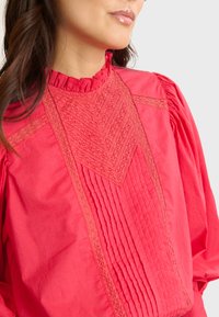 Blusa rosa con un alto cuello con volantes, elegante encaje detallado y plisados verticales en la parte frontal para añadir textura y contraste en el diseño.