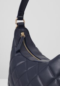 Marinblå quiltad skinnhandväska med gulddragkedja och mjuk yta. Har en bekväm axelrem och rundad form.