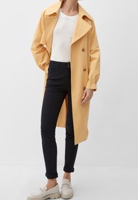 Geel lichtgewicht trenchcoat met een grote kraag en dubbele knopen, gecombineerd met marineblauwe skinny jeans en beige instappers.