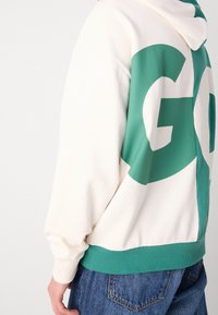 Krémově zbarvená mikina s kapucí s velkou zelenou grafikou "GO" na zádech, žebrovaným lemem a manžetami a kapucí s nastavovacím páskem. Měkká, hladká textura.