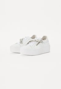 Hvide sneakers med en tyk, relieffsål. Har en skinnende sølvkæde detalje over det øverste og en lynlås på siden som designelement.