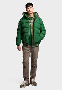 Green puffer jacket with a hood, featuring buttons and a patch, layered over a plaid shirt. Beige pants and multicolored sneakers complete the look.