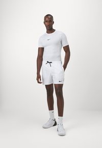 Camiseta deportiva blanca de manga corta con un logotipo negro de Nike, combinada con pantalones cortos blancos con cordón y detalles laterales, llevada con zapatillas blancas.