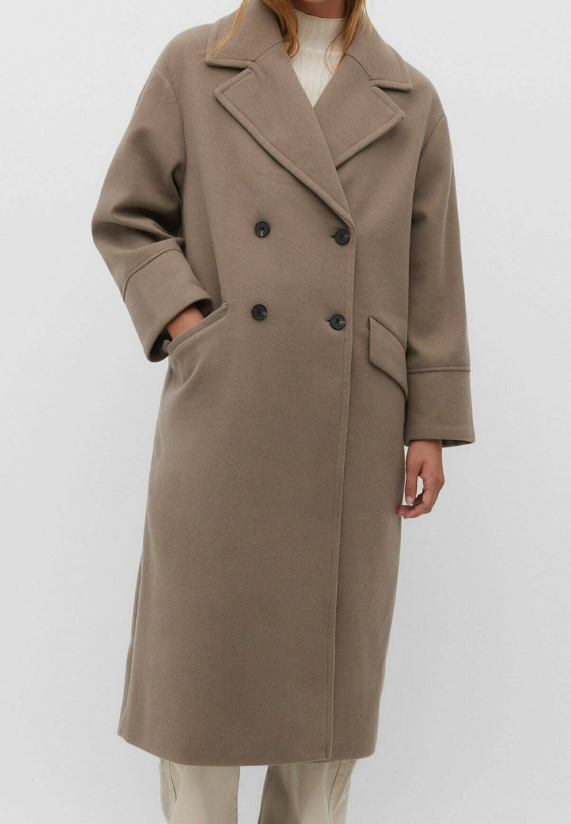 Femme portant un long manteau en laine taupe à double boutonnage avec des boutons noirs, de larges revers et des poches latérales, associé à un pantalon beige clair.