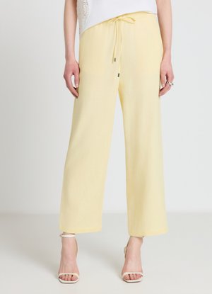 Woman wearing pale yellow wide-leg pants with drawstring waist and white open-toe ankle-strap heels, standing against plain light background.