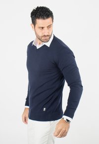Pull en V marine, en matière tricotée, coupe ajustée, avec des poignets et un ourlet à fines côtes, portant une chemise blanche en dessous, avec un petit logo.