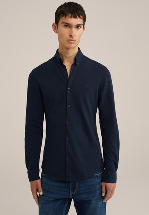 Camisa de botones azul marino con mangas largas, que presenta un cuello extendido y un sutil logotipo en el pecho. Fabricada con un tejido suave.