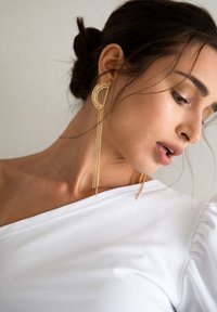 Gouden oorbellen met een geometrische maansikkelvorm, versierd met kettingen die vanaf de onderkant hangen. Het model heeft donker haar dat in een lage knot is gestyled.