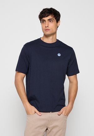 Giovane uomo che indossa una t-shirt blu navy tinta unita a maniche corte con un piccolo logo rotondo sul petto, in piedi con le mani nelle tasche di pantaloni beige.