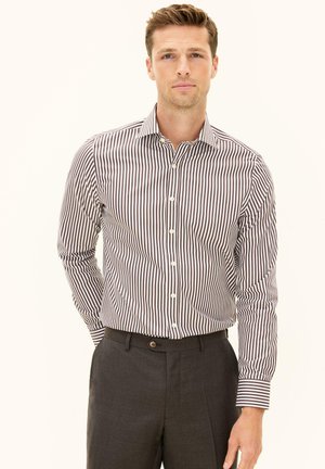 Mężczyzna w brązowo-białej koszuli w pionowe paski, włożonej do ciemnobrązowych spodni, stojący na jasnym tle.