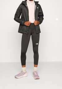 Veste noire légère avec capuche, intérieur gris. Haut rose à col zippé. Leggings sombres avec logo. Baskets roses avec détails texturés et semelles blanches.