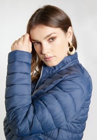 Giacca piumino blu navy con texture trapuntata e colletto alto; presenta una chiusura frontale con zipper e una finitura liscia e lucida. Orecchini rotondi dorati.