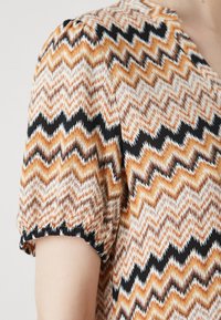 Μπλούζα κοντού μανικιού με ζιγκ-ζαγκ σχέδιο σε μαύρο, πορτοκαλί, καφέ και κρέμ χρώμα; διαθέτει ελαστικούς σφιγκτήρες και ελαφριά υφή υφάσματος.