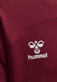 Burgundyröd tröja med en texturerad yta har en vit logotyp av en bi och varumärket "hummel" tydligt utställd.