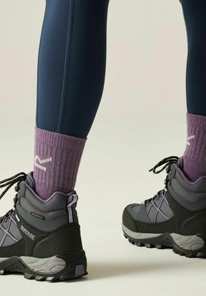 Persona con calcetines morados y botas de senderismo impermeables grises con suelas y cordones negros, de pie sobre un fondo neutro.