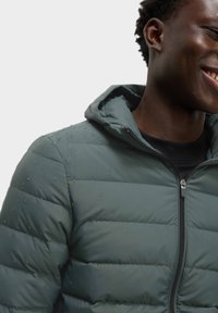 Groene pufferjack met een ritssluiting en een capuchon. Waterdruppels zijn zichtbaar op het oppervlak, wat het weerbestendige materiaal en de textuur benadrukt.