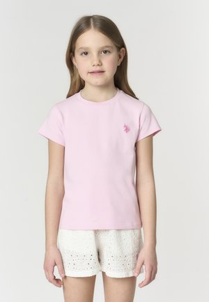 Ragazza giovane con capelli lisci che indossa una maglietta a maniche corte rosa chiaro e pantaloncini bianchi con motivo a occhiello, in piedi contro uno sfondo semplice.