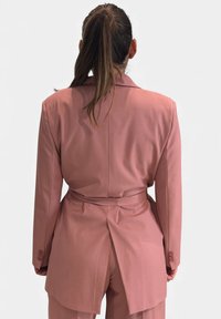 Blazer rosa cipria su misura con vita con cintura, spacco singolo sul retro e maniche lunghe. Tessuto morbido con texture liscia.