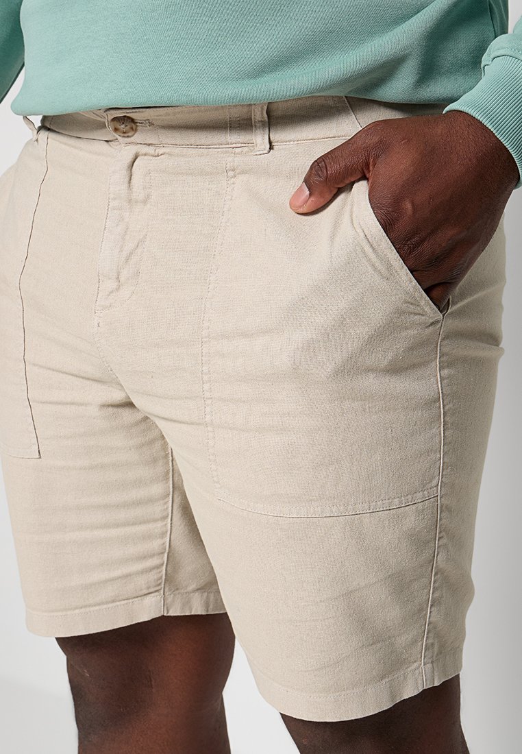 Beżowe lniane szorty w luźnym kroju, z kieszeniami po bokach i złożonym wykończeniem. Nosi się je z miętowozieloną bluzką, prezentując swobodny wygląd.