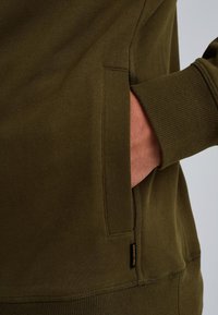 Felpa verde oliva con tessuto strutturato, dotata di tasca laterale e polsini a coste. Etichetta visibile sulla cucitura laterale.