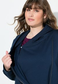 Chaqueta de marina con una tela suave y texturizada, que presenta un cuello chal, cremallera frontal y un cierre de botón. Llevada sobre una blusa color burdeos.