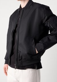 Veste bomber noire avec une finition lisse et brillante, poignets et ourlet côtelés. Comprend des poches latérales et une fermeture éclair avant avec un curseur distinctif.