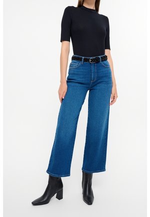 Frau trägt ein schwarzes, eng anliegendes Oberteil, hoch taillierte, weit geschnittene blaue Jeans mit schwarzem Gürtel und schwarze Stiefeletten vor weißem Hintergrund.