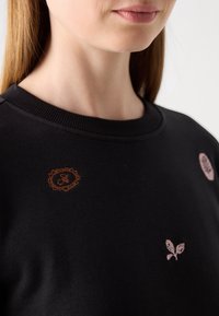 Gros plan d'une personne portant un pull noir avec de petits motifs floraux et circulaires brodés près de l'encolure.