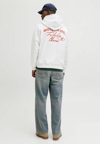 Pelēks kokvilnas kapučinos džemperis ar lielu sarkanu grafiku uz aizmugures, apvienots ar brīviem ziliem džinsu biksēm un brūnu apavu.