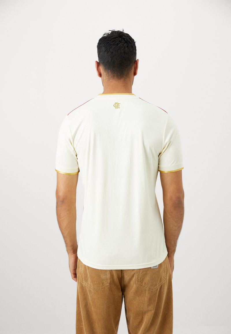 Kurzärmliges cremefarbenes Sporttrikot mit roten Schulterakzenten und goldenen Zierleisten. Verfügt über ein kleines Logo am oberen Rücken und einen strukturierten Stoffdetails.