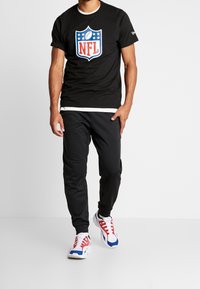 Hombre vestido con una camiseta negra con el logo de la NFL, pantalones jogger negros y zapatillas blancas con detalles rojos y azules, de pie contra un fondo blanco.