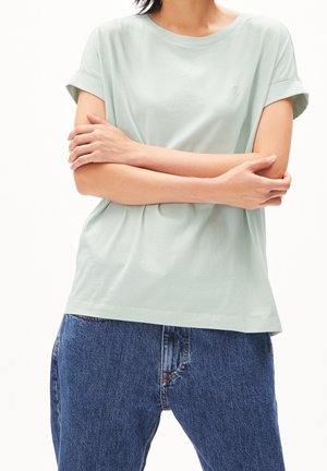 T-shirt en coton vert clair avec manches retroussées et col rond, porté avec un jean denim bleu. Détail subtil du logo sur la poitrine.