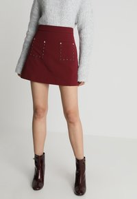 Mini-jupe ajustée bordeaux avec des accents d'étoiles argentées et des détails de œillets décoratifs sur les poches, associée à un pull gris clair et des bottes brillantes.